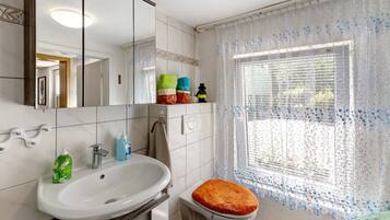A bathroom with a white sink, mirror, and toilet. There are toiletries on the counter and a window with a patterned curtain.