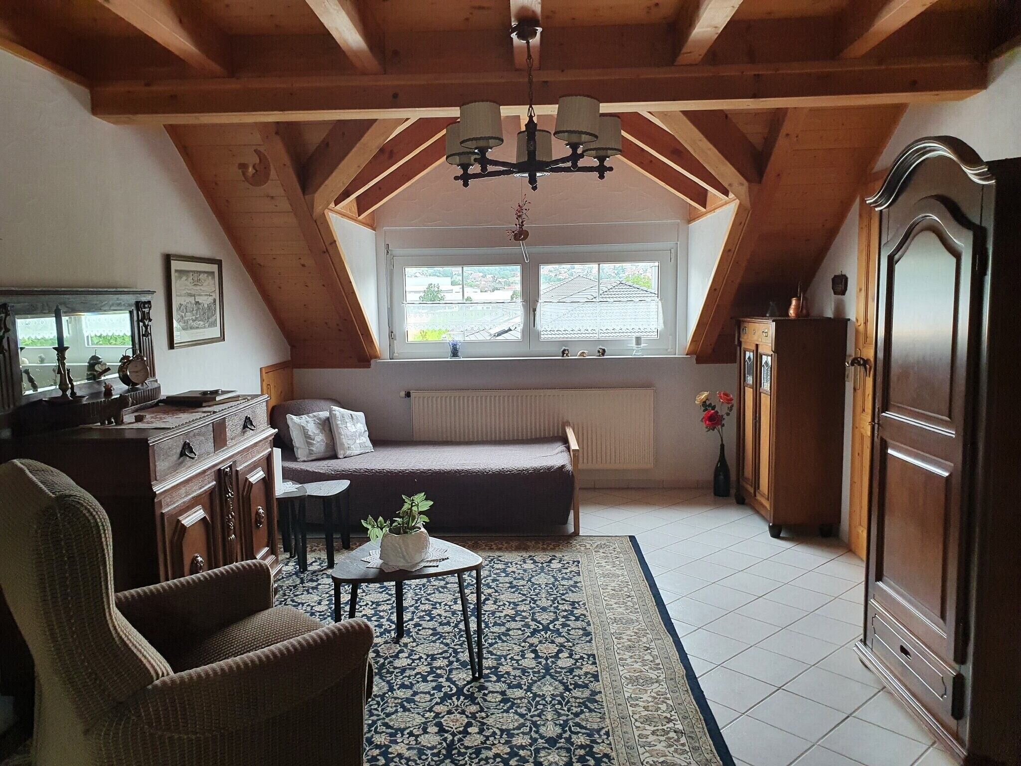 Une chambre mansardée chaleureuse, avec un plafond en pente, une armoire en bois, un radiateur, une petite table avec une plante et une fenêtre donnant sur l’extérieur.