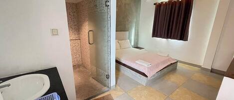 A hotel room with a shower, a bed, a bathroom with a sink and a basket, and a window with curtains.