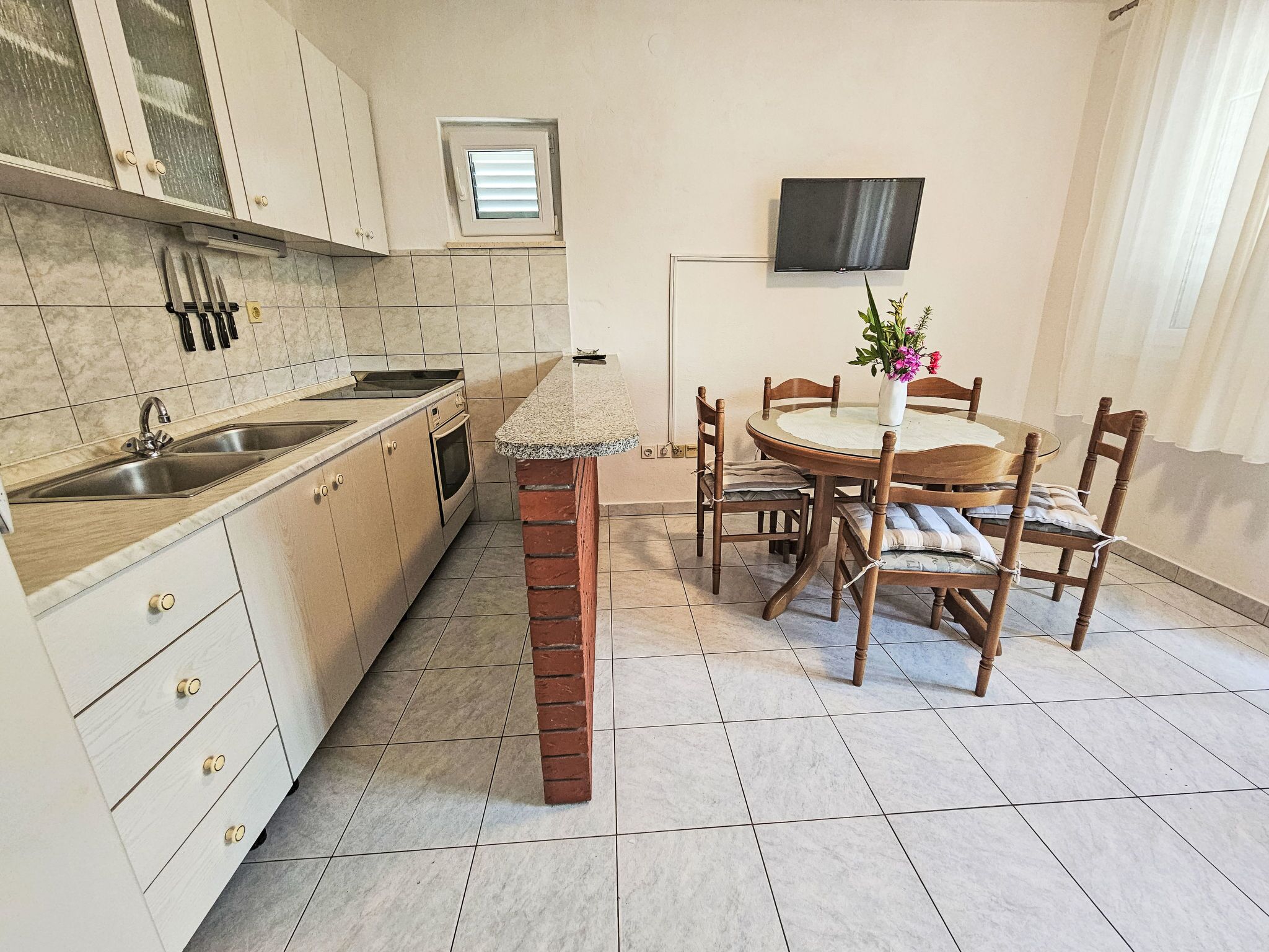 A compact kitchen with a sink, stove, and cabinets, a dining table with chairs, and a TV mounted on the wall.