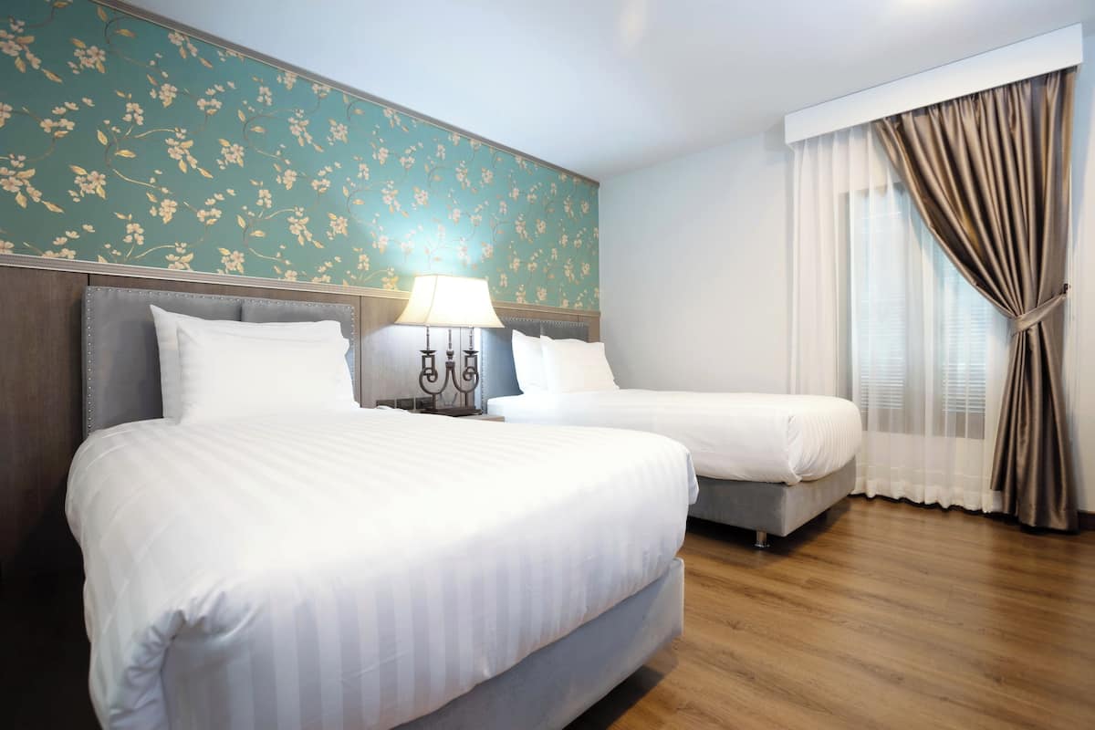 A hotel room with two beds, a nightstand with a lamp, floral wallpaper, and a window with curtains.