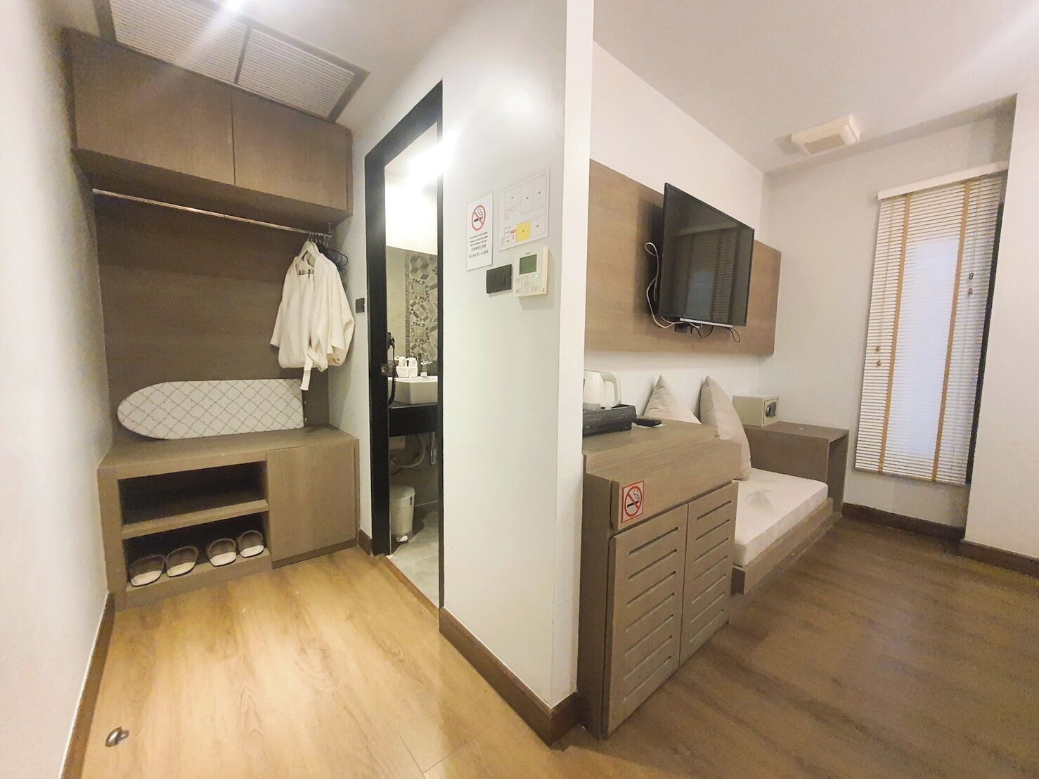 A compact bedroom with a built-in wardrobe, a television mounted on the wall, and a window with blinds.