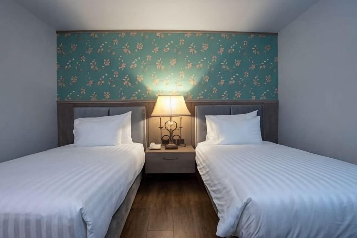 A twin room with two beds, a nightstand with a lamp, and floral wallpaper.