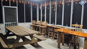 An outdoor patio area with picnic tables and chairs, a ceiling fan, string lights, and large windows.