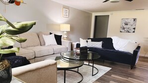 A living room with a beige sofa, a dark blue sofa, a glass coffee table, and a small side table with a lamp.