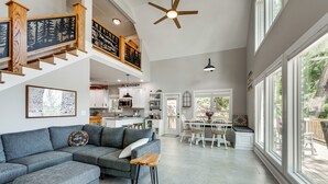 A spacious living area with a grey sectional sofa, a wooden bench, and a dining table with chairs. A kitchen is visible in the background, and there are large windows providing natural light. - Brand-New Perry House w/ Kayaks & Lake Access (Perry)