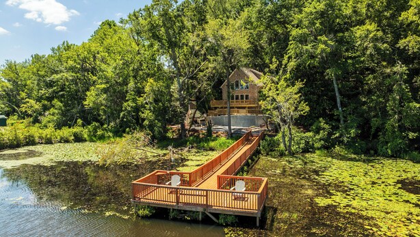 A wooden deck with two chairs extending over a pond, surrounded by dense greenery and a cabin in the background. - Brand-New Perry House w/ Kayaks & Lake Access (Perry)