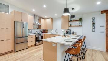 A modern kitchen with wooden cabinets, a stainless steel refrigerator, a central island with bar stools, and pendant lighting.