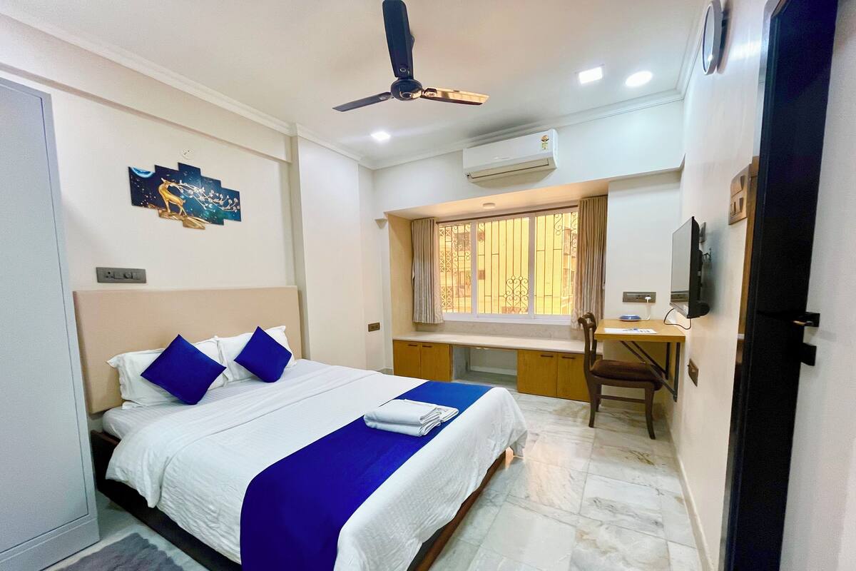 A hotel room with a bed, a desk, a chair, a television, and an air conditioning unit.