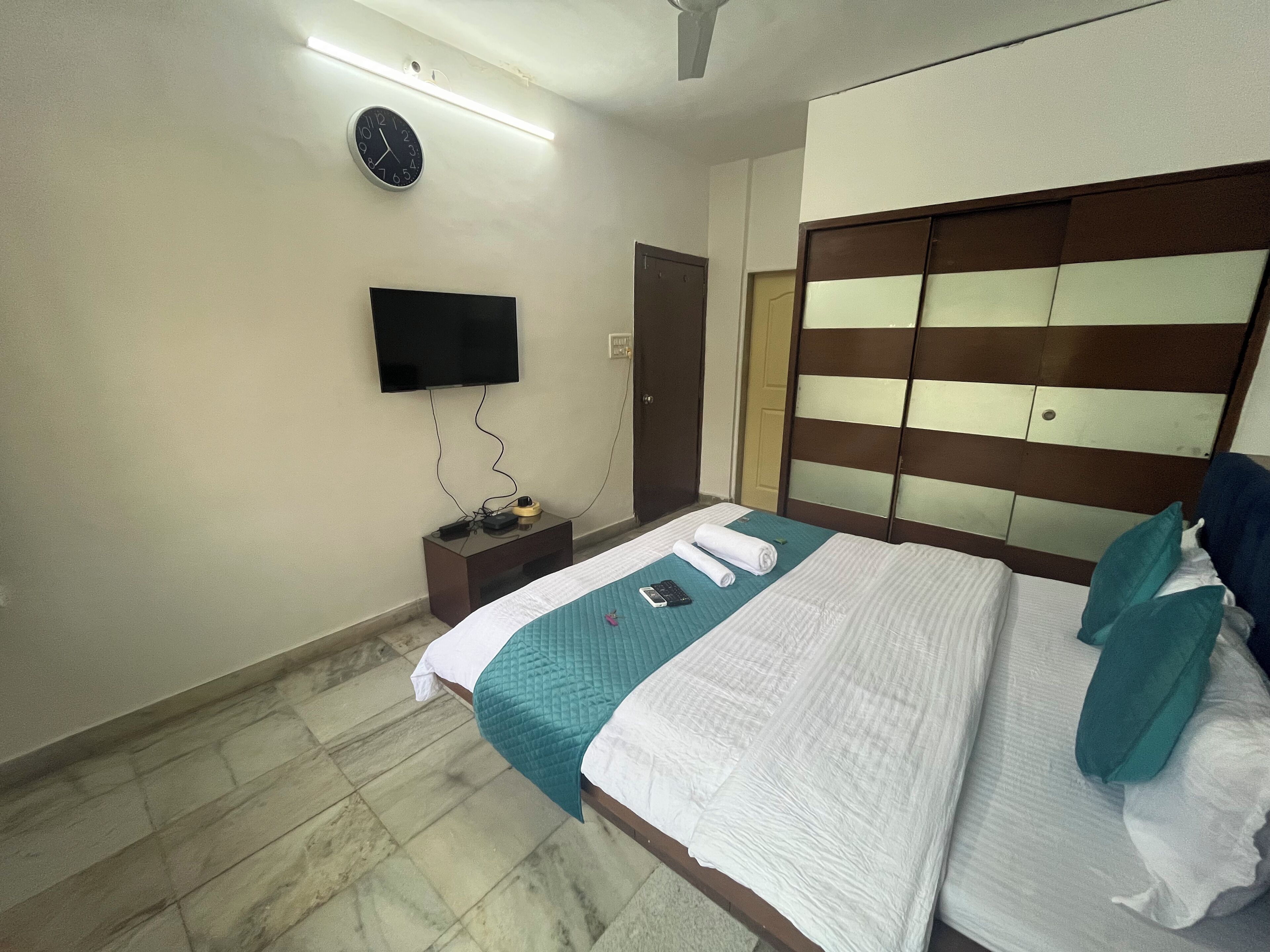 A hotel room with a bed, a TV mounted on the wall, a nightstand with a phone and charger, and a wooden wardrobe.