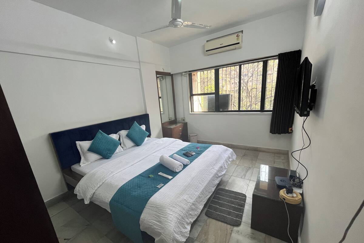 A hotel room with a bed, a ceiling fan, an air conditioning unit, a TV, and a window with curtains.