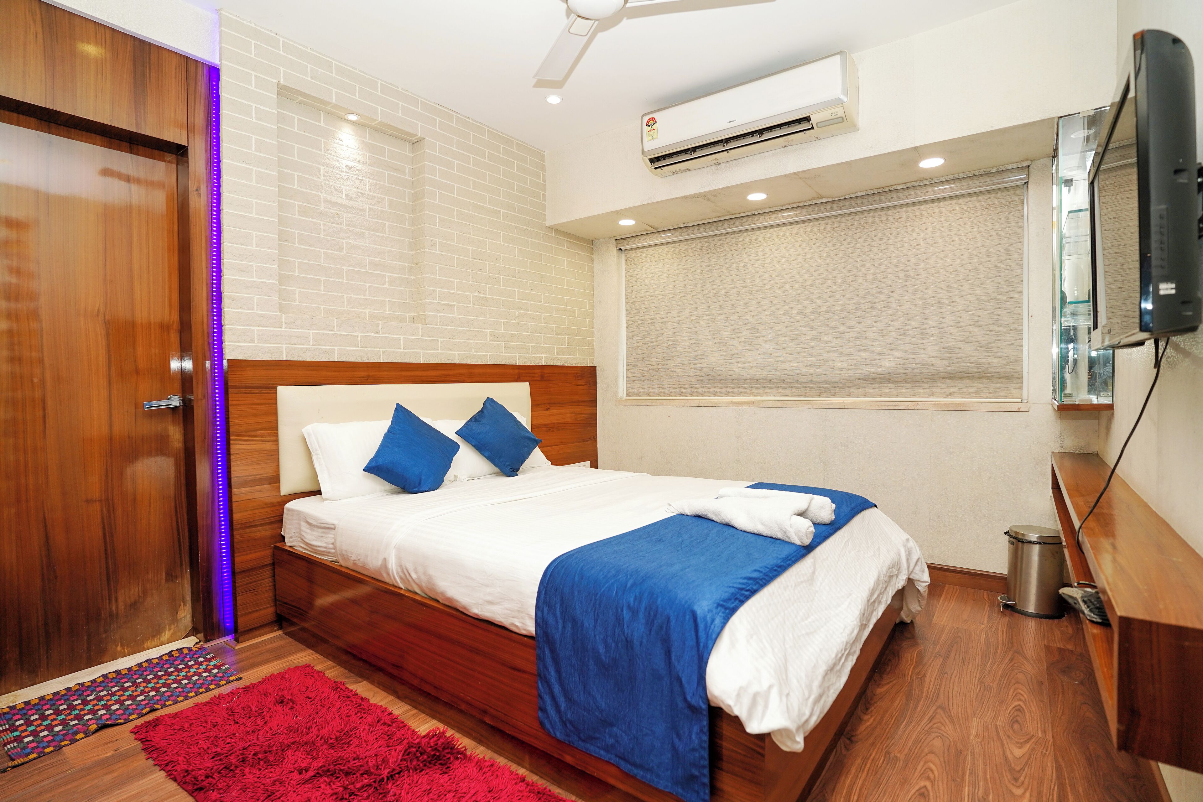 Quarto bem arrumado com cama de madeira, almofadas azuis, tapete vermelho e unidade de ar condicionado branca na parede.