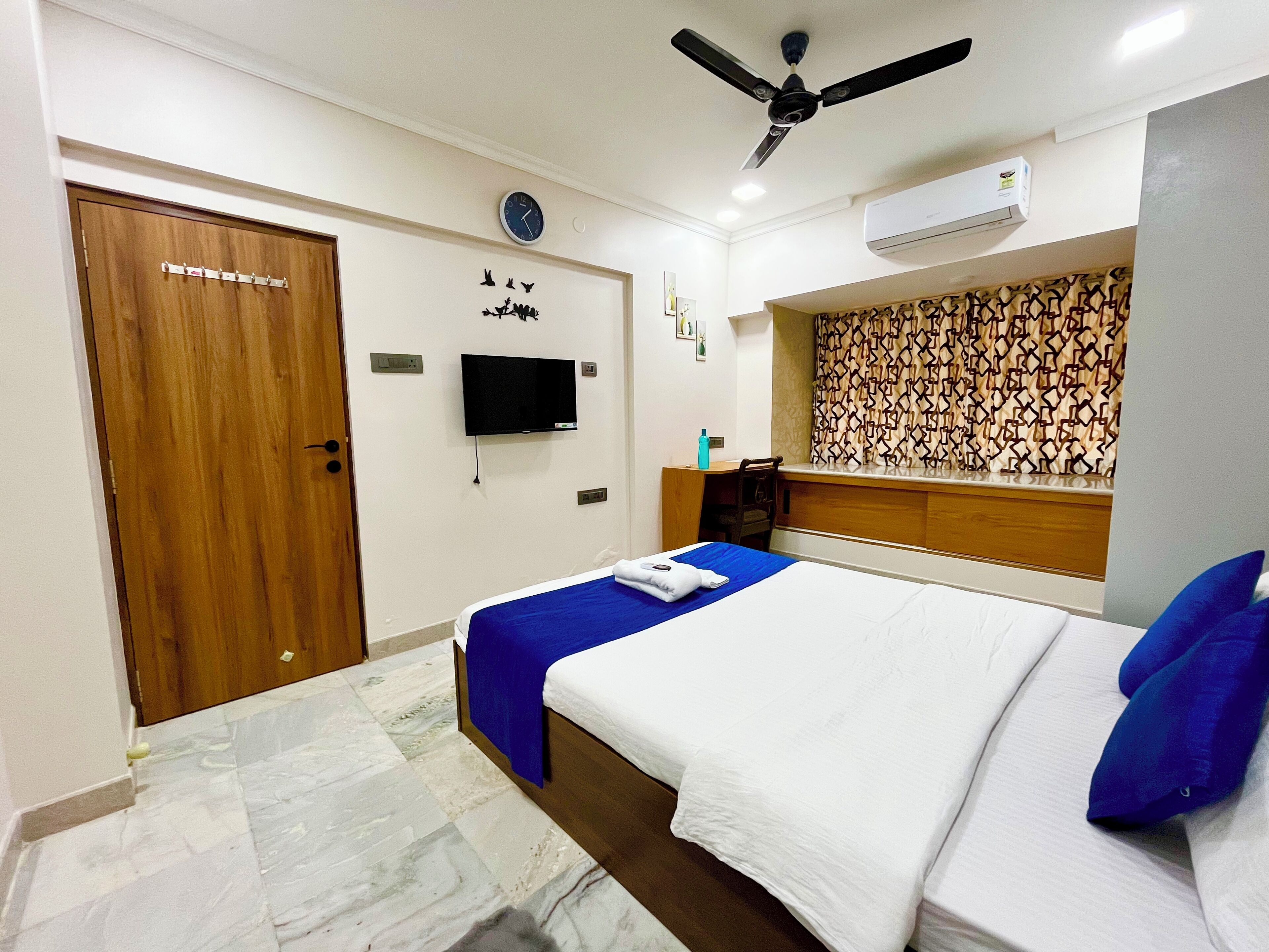 Une chambre d’hôtel équipée d’un lit, d’un bureau, d’une télévision, d’un ventilateur de plafond et d’un rideau à motifs.