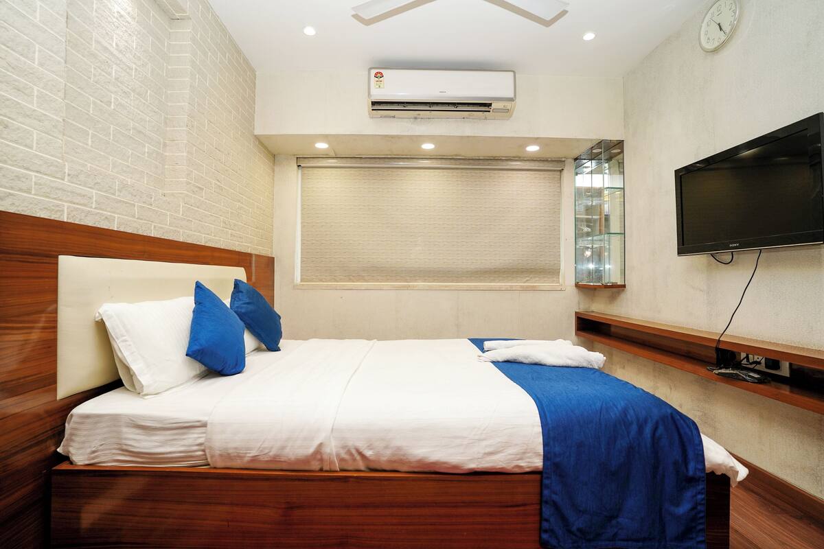 A hotel room with a bed, a TV, an air conditioning unit, and a window with blinds.