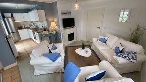 Uma sala de estar moderna com um sofá branco, uma televisão, uma lareira e uma cozinha ao fundo.