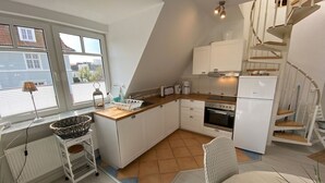 A compact kitchen with white cabinets, a stove, and a sink. There is a window with blinds, a radiator, and a small table with a basket. - Apartment 'Ferienwohnung Parkblick 2' with Private Terrace, Balcony and Wi-Fi (Lübeck Travemünde)