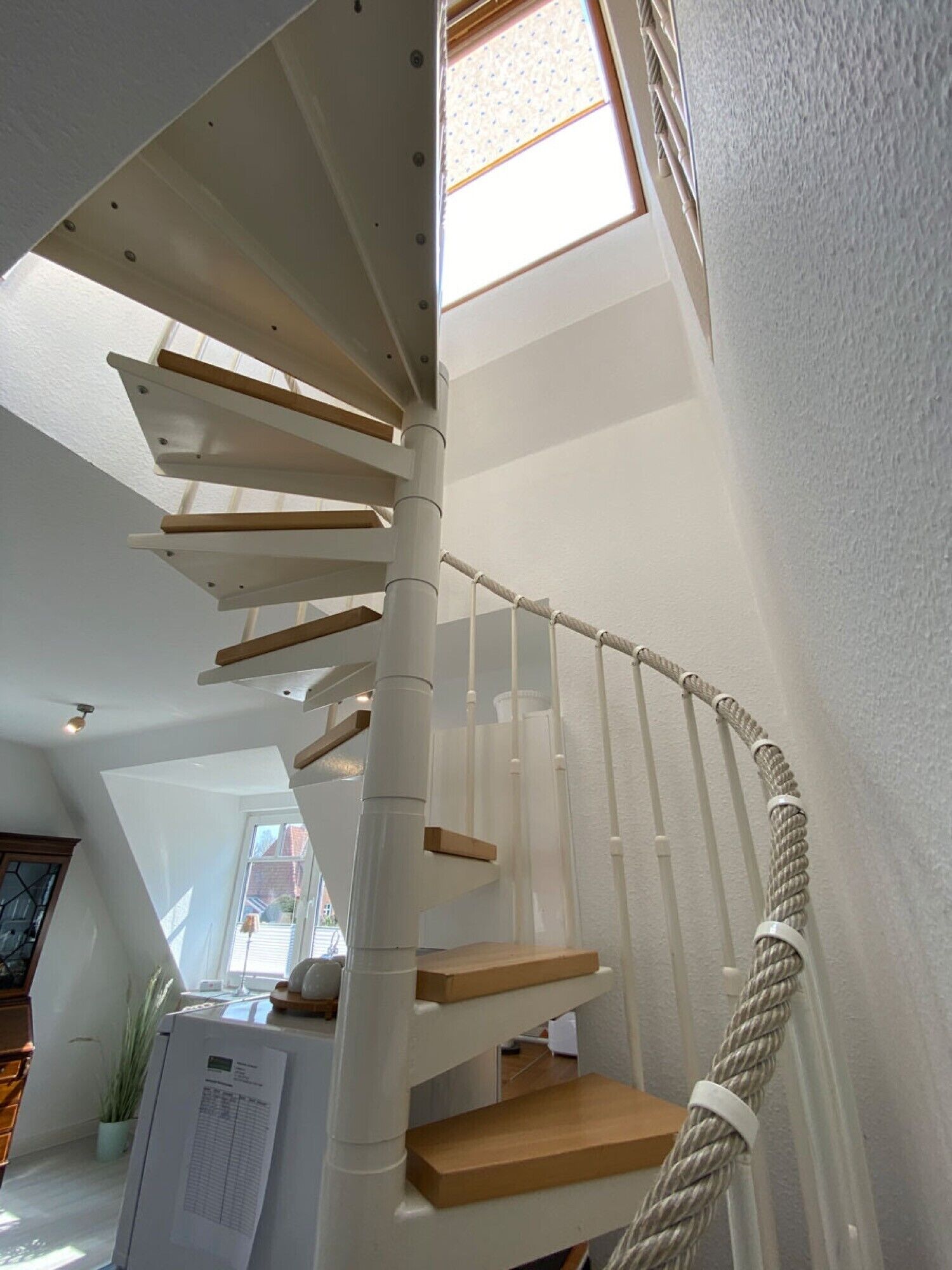 Un escalier en colimaçon avec des marches en bois et une main courante en corde, menant à une lucarne.