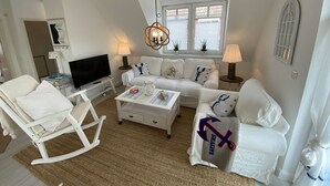 A living room with a TV, white furniture, a chandelier, and a window with blinds. - Apartment 'Ferienwohnung Parkblick 2' with Private Terrace, Balcony and Wi-Fi (Lübeck Travemünde)