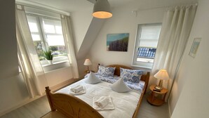 A bedroom with a wooden bed, two bedside tables, a window with curtains, a painting on the wall, and a ceiling light. - Apartment 'Ferienwohnung Parkblick 2' with Private Terrace, Balcony and Wi-Fi (Lübeck Travemünde)