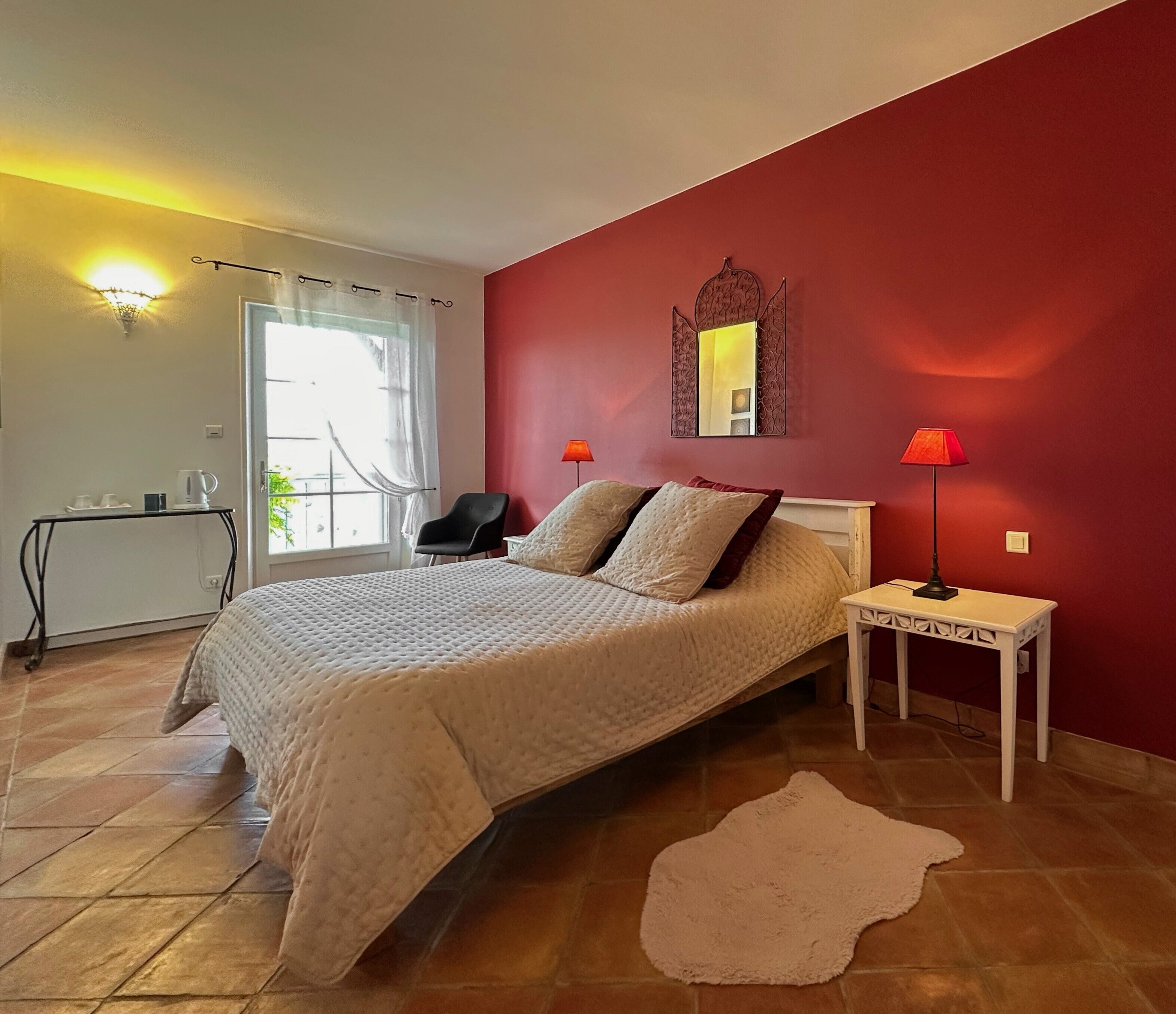 A bedroom with a bed, a nightstand, a chair, a table, a mirror, and a window with curtains.