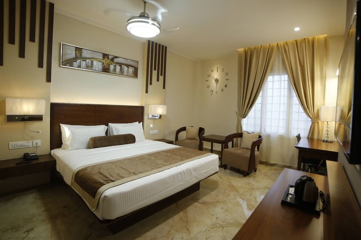 A hotel room with a large bed, bedside tables, a desk, and a chair. There is a clock on the wall and a window with curtains.