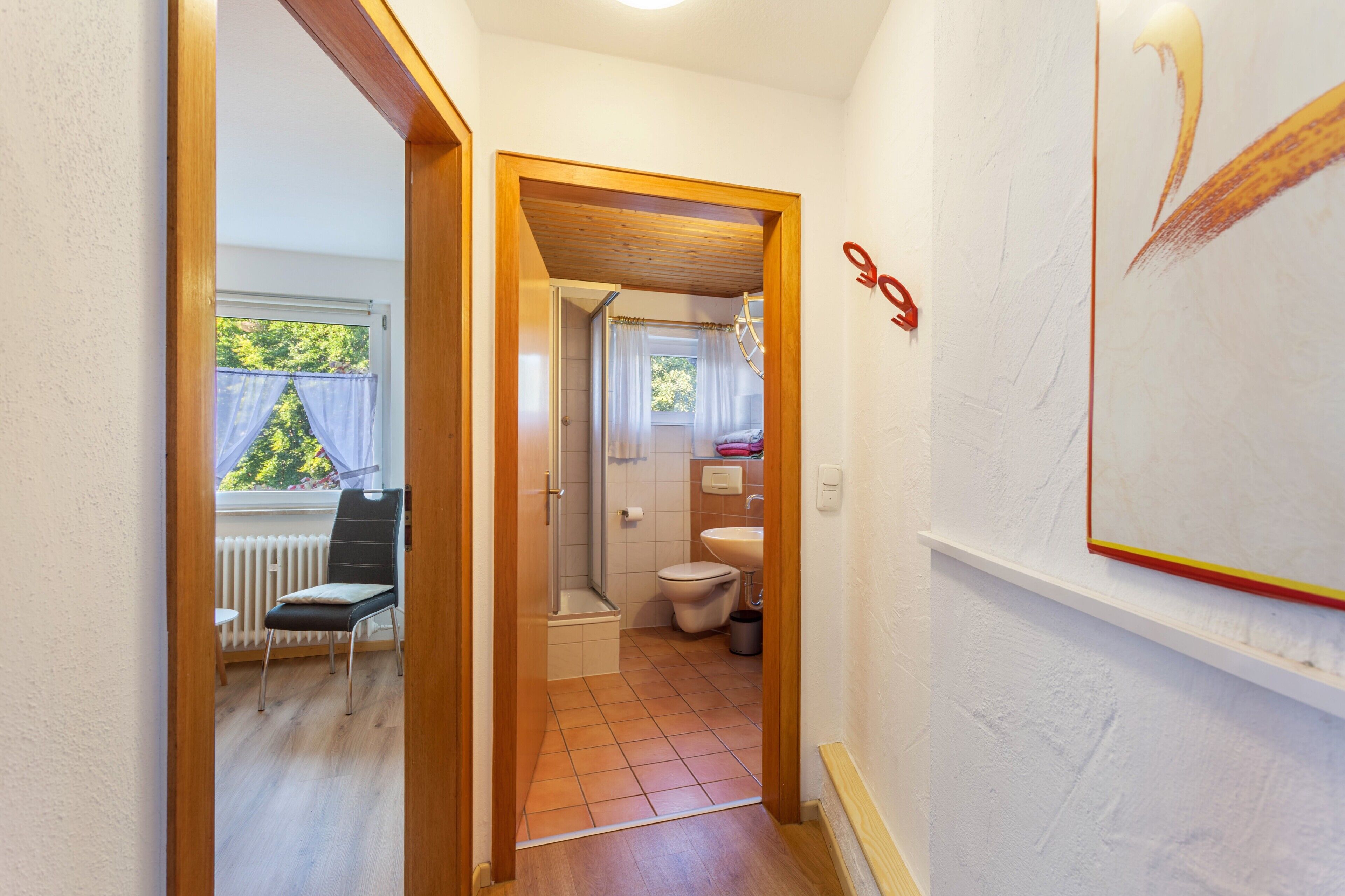 Un couloir étroit mène à une salle de bain équipée d’une douche, de toilettes et d’une chaise placée près d’une fenêtre.