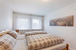 Ein Schlafzimmer mit zwei Betten, einem Fenster mit durchsichtigen Vorhängen, einem Nachttisch mit Lampe und einem Bild eines Stiers an der Wand.