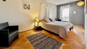 A bedroom with a bed, a nightstand, a chair, a window with curtains, and a hanging plant. - Maison Oléa - Chambre D'hôtes "naturéa" Avec vue sur la Vallée de la Vézère (Le Bugue)