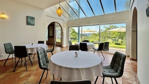 A dining area with round tables, chairs, and a view of greenery through large windows. - Maison Oléa - Chambre D'hôtes "naturéa" Avec vue sur la Vallée de la Vézère (Le Bugue)