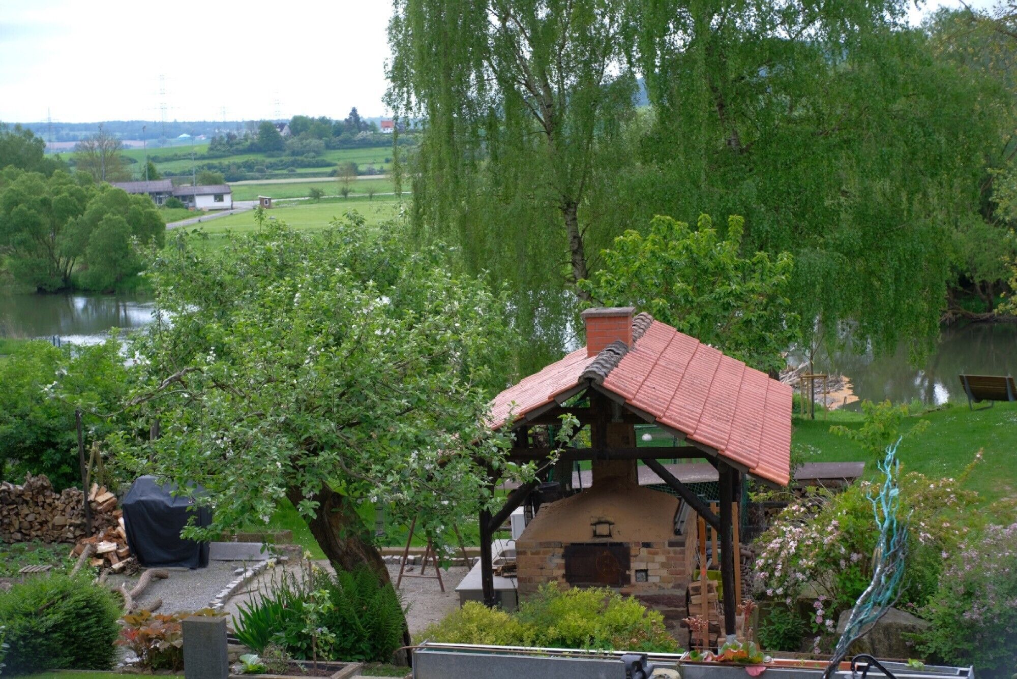 Un espace extérieur aménagé dans un style rustique, avec une structure en briques, entouré d’une végétation luxuriante et offrant une vue sur une rivière.