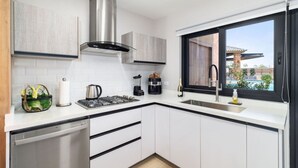 A modern kitchen with white cabinetry, a stainless steel range hood, a built-in dishwasher, and a window with a view of a pool and buildings. - LUX 3BR House Pool Gym 7-Min Walk Palm Beach - Porta Al Sole 7 by Bocobay (Noord)