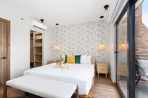 Une chambre moderne avec un grand lit, des tables de chevet, un banc et une porte-fenêtre coulissante donnant sur un balcon.