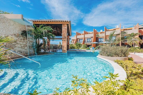 Una piscina con caída de agua, rodeada por una zona de piscina con sillas y mesas.