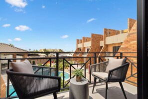 Ein Balkon mit zwei Korbstühlen, einem kleinen Tisch mit einer Pflanze und Blick auf einen Swimmingpool sowie Gebäude.