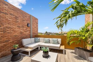 Eine Dachterrasse mit Ziegelwand, einem weißen Sofa mit Kissen, einem kleinen Tisch und Topfpflanzen.