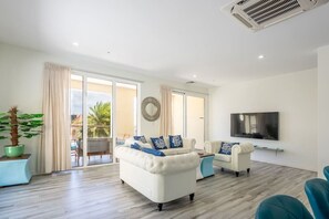 Una sala de estar moderna con un sofá blanco, un sillón blanco más pequeño y un sillón azul. Hay un televisor de pantalla plana grande montado en la pared, una planta en maceta y un ventanal corredero que da a un área exterior.