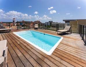 Terraza de madera con un jacuzzi rectangular, sillas de descanso y vista despejada de los edificios circundantes.