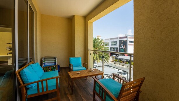 Un balcón con mobiliario de madera, que incluye una mesa y dos sillas, y vistas a un paisaje urbano con edificios y palmeras.