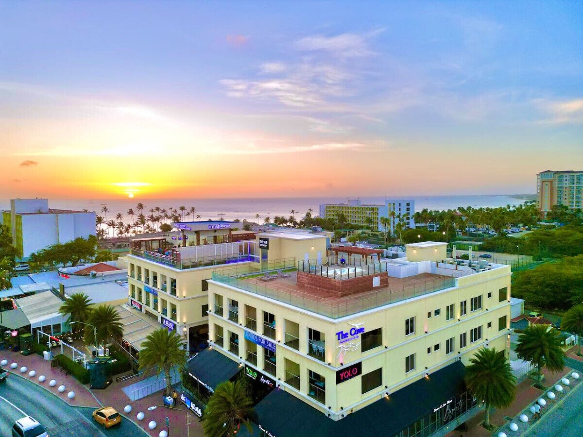 Un hôtel à plusieurs étages doté d’un bar sur le toit, situé près d’une plage bordée de palmiers, avec un coucher de soleil en toile de fond.
