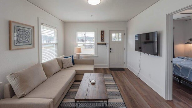 A modern living room with a beige sofa, a coffee table, a TV, and a wooden floor. - #StayInMyDistrict Bluebird Cottage Coos Bay (Coos Bay)