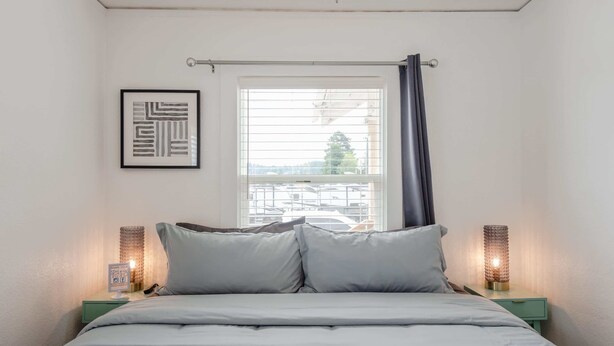 A bedroom with a bed, a window with blinds, a framed picture on the wall, and bedside lamps.