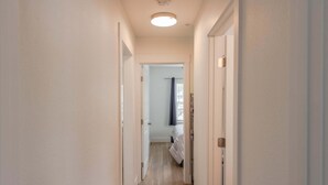 Un couloir étroit aux murs blancs et au plancher en bois mène à une chambre à coucher équipée d’un lit et d’une fenêtre avec des rideaux.