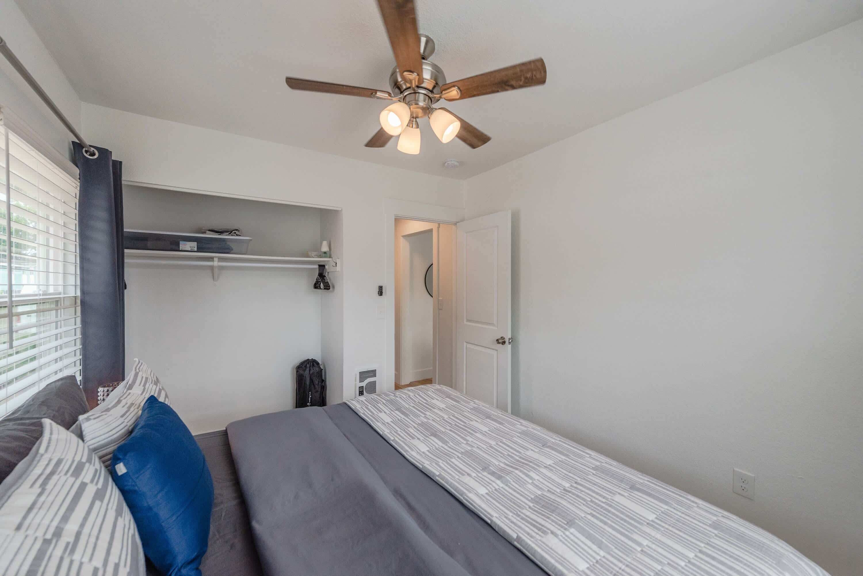 Un dormitorio con cama, ventilador de techo, ventana con persianas, puerta y estante con objetos.