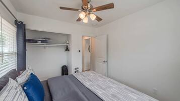 Un dormitorio con cama, ventilador de techo, ventana con persianas, puerta y estante con objetos.