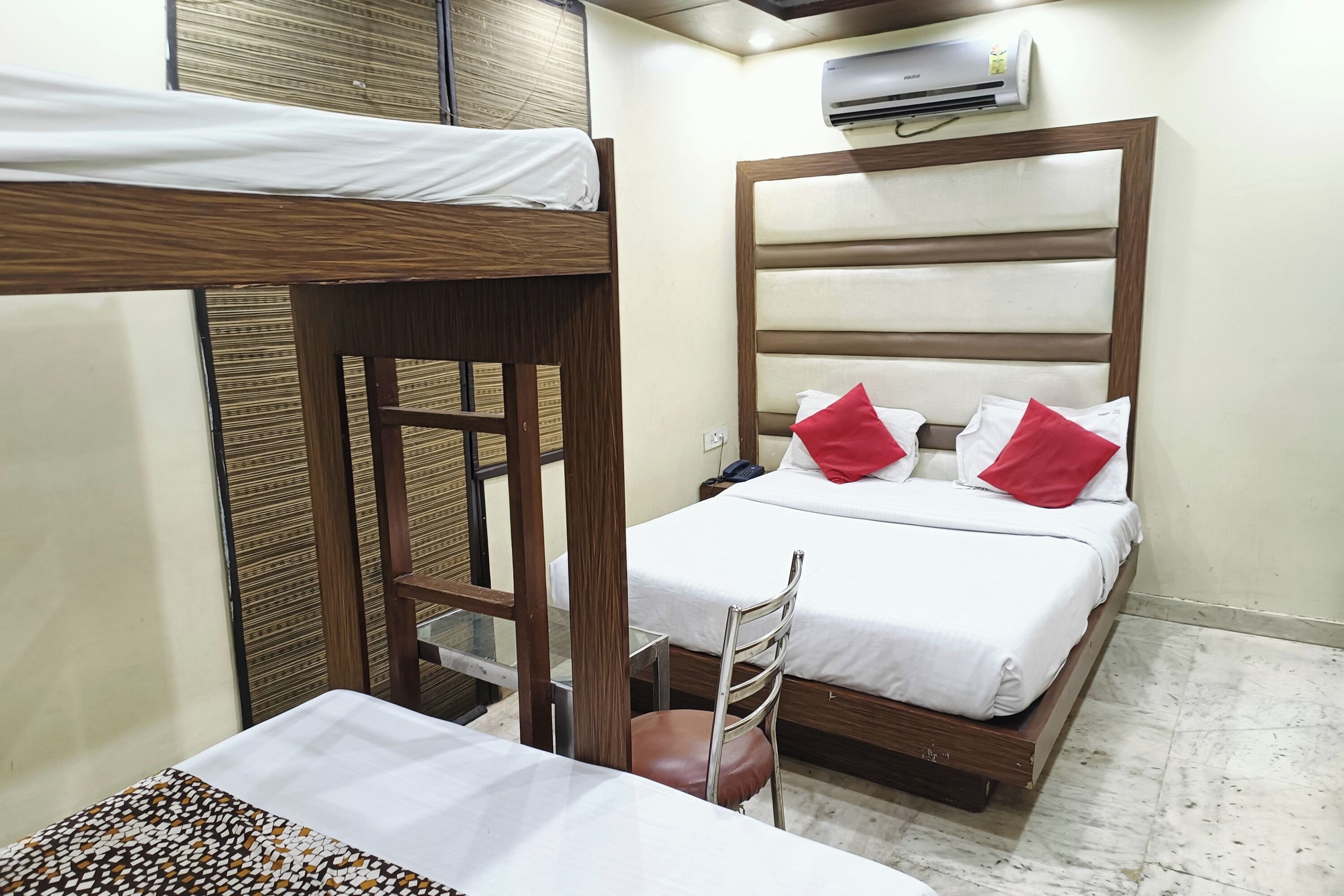 A room with a bunk bed, a single bed with red pillows, a chair, and an air conditioning unit on the wall.