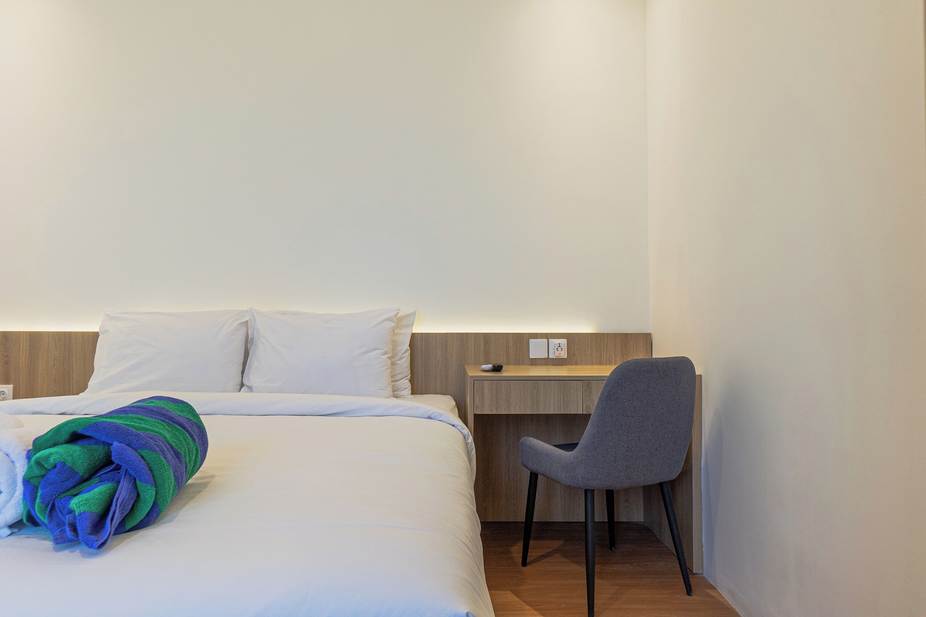A neatly made bed with a striped blanket, a grey chair, and a wooden desk in a hotel room.