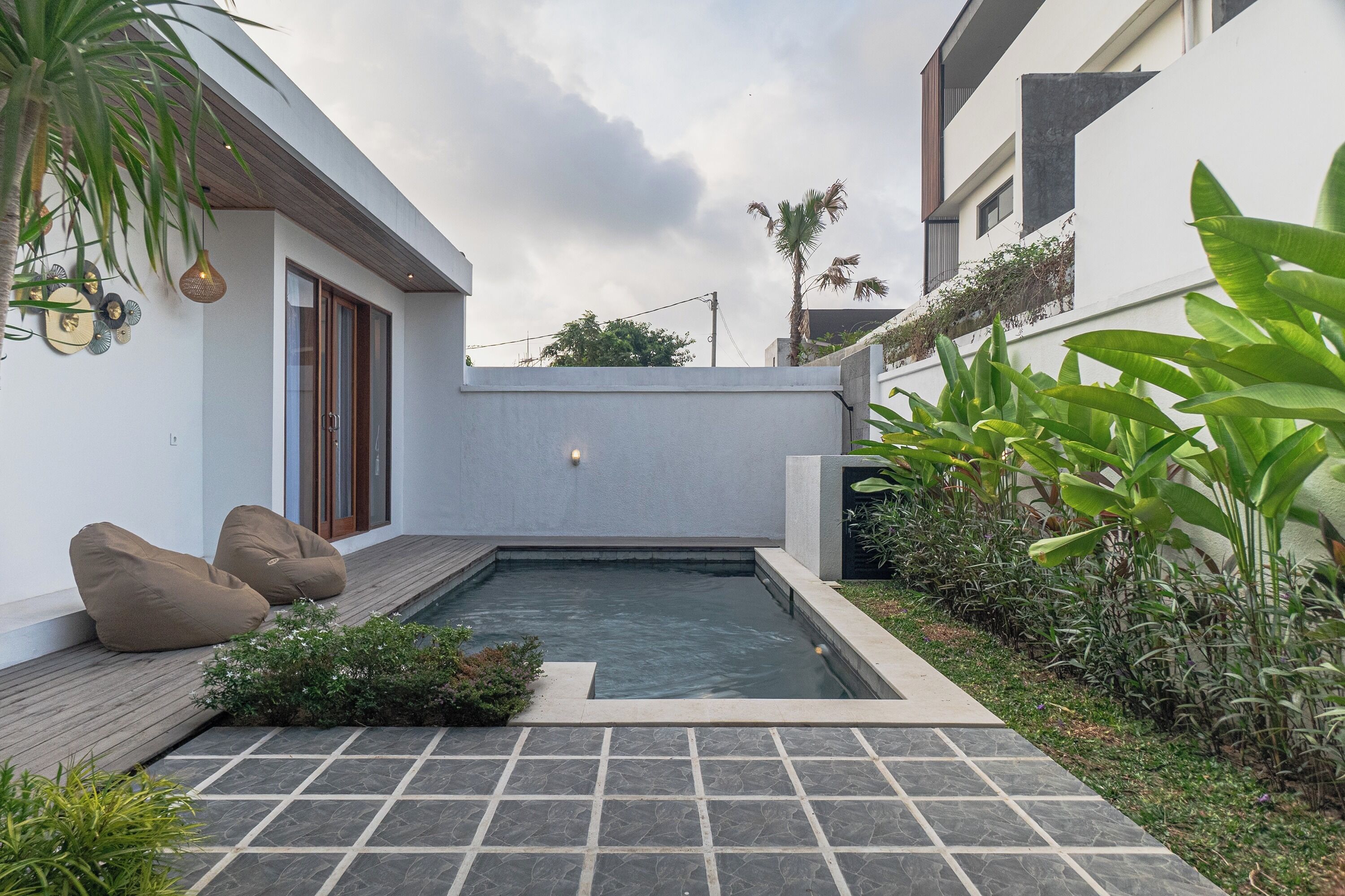 A modern outdoor pool area with a wooden deck, seating, and lush greenery.