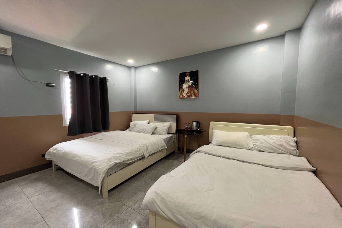 Ein Hotelzimmer mit zwei Betten, einem kleinen Tisch, einem Wandbild und einem Fenster mit Vorhängen.