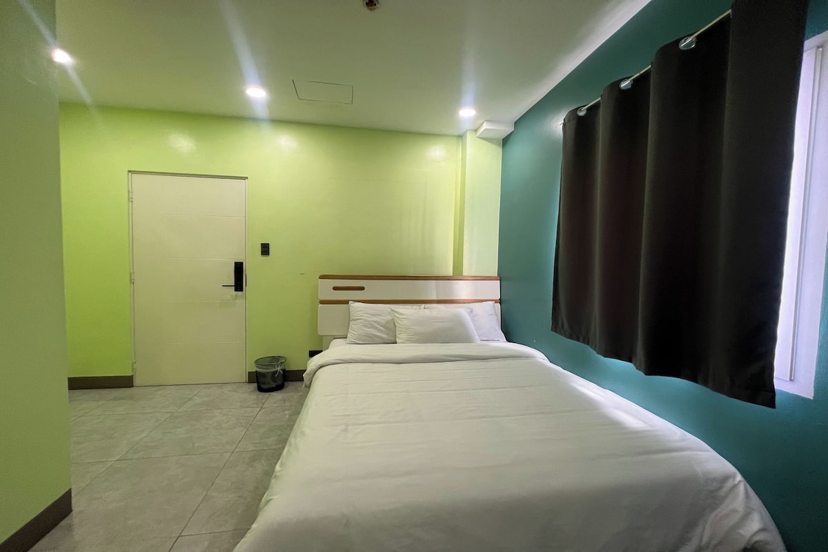 Ein Hotelzimmer mit Bett, Tür, Mülleimer und einem Fenster mit Vorhängen.