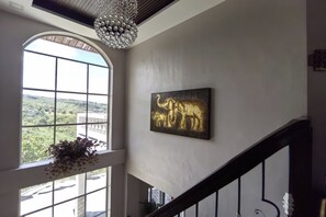 A spacious room with a large arched window offering a view of greenery outside. A chandelier hangs from the ceiling, and a framed artwork of elephants is displayed on the wall. - Tampus Staycation Rental Cebu (Talisay)